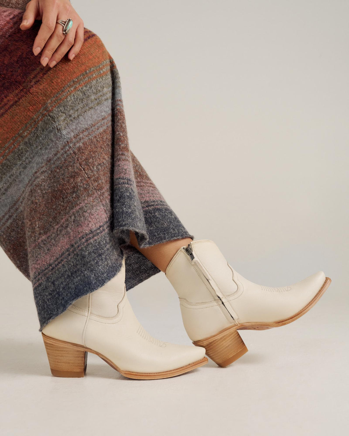 A person wearing cream-colored, mid-calf heeled cowboy boots and a multicolored, textured skirt standing on a light surface.