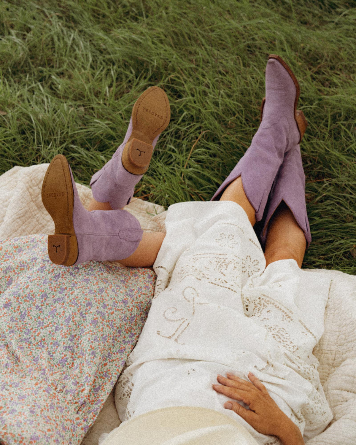 The Annie in Lavender Suede. Two people lie on a blanket in the grass, wearing light-colored skirts and stylish lavender suede cowboy boots, their legs extended and the boots beautifully visible.