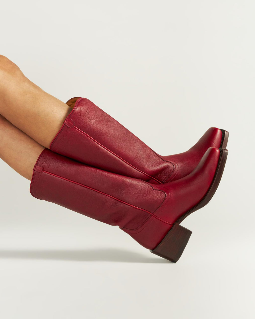 The Charlie in Scarlet Cowhide. A person wearing tall, scarlet red leather cowboy boots with block heels—crafted in the signature Charlie Cowhide style—legs extended sideways against a plain white background.