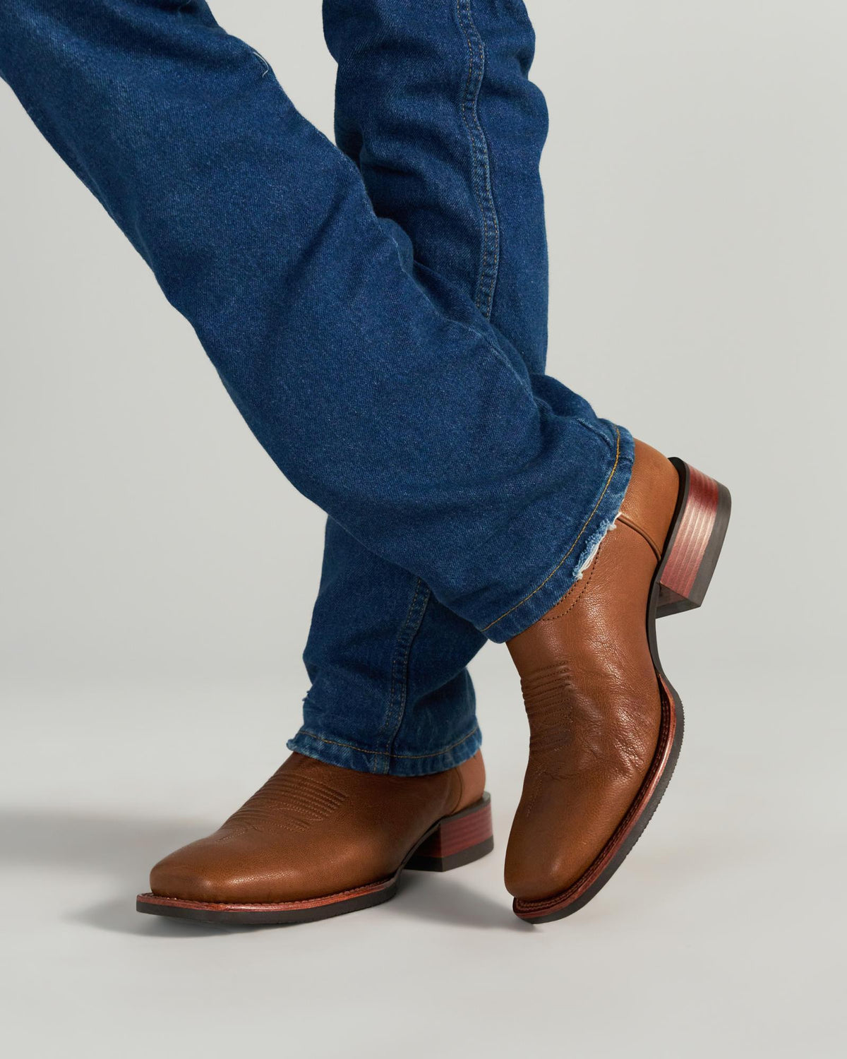 The Dusty in Mesa Goat. Person wearing blue jeans and brown leather cowboy boots stands with legs crossed on a plain background, evoking the rugged spirit of The Dusty Goat.