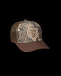 A brown and camouflage trucker hat with a mesh back and a round patch reading “Tecovas Mossy Oak” on the front.