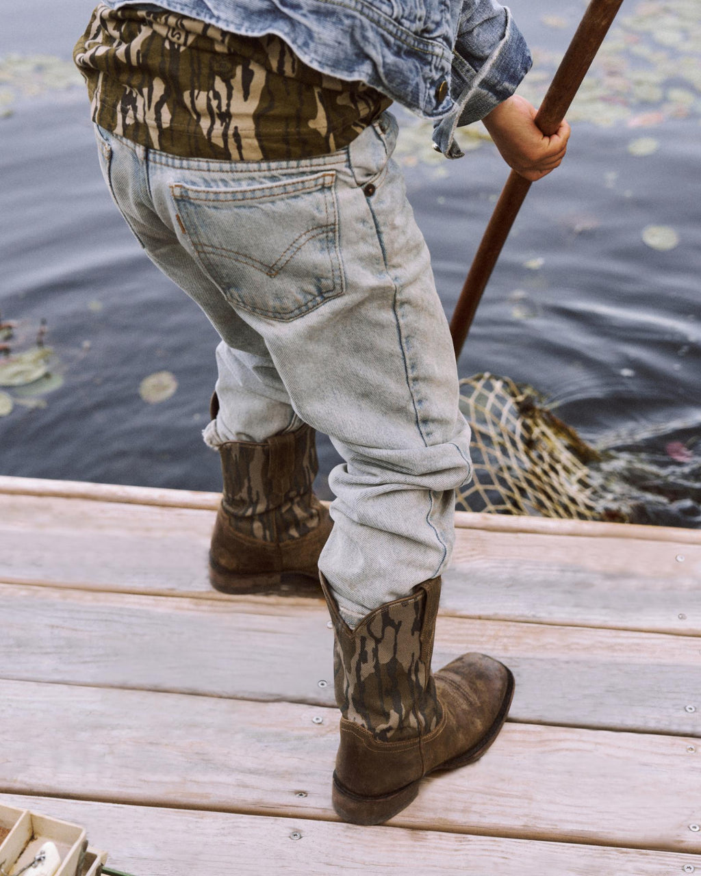 Person wearing denim jeans and camouflage boots uses a fishing net at the edge of a wooden dock over water with lily pads.