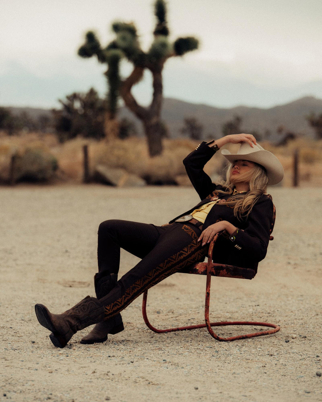 A person in a cowboy hat and western suit sits on a rusted chair in a desert landscape with Joshua trees behind, gazing thoughtfully as The Tess Goat cafe sign peeks from the sun-baked distance.