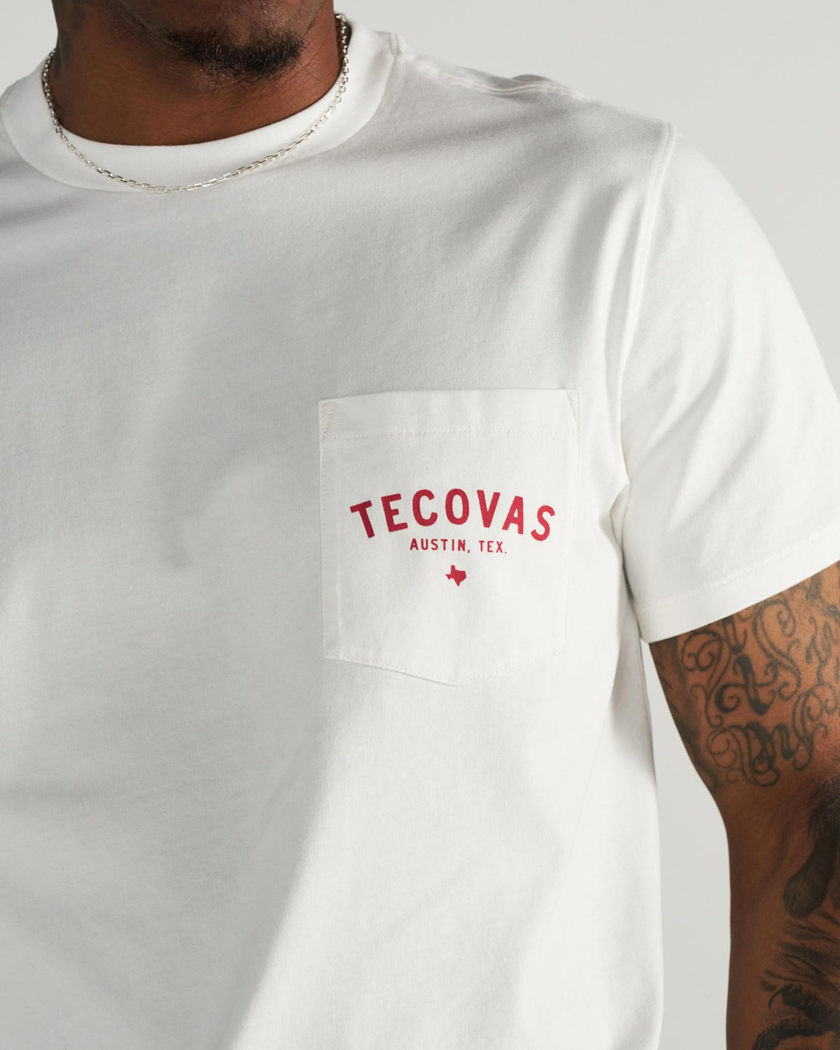 Man wearing a white T-shirt with “Tecovas Austin, Tex.” in red letters on the front pocket. Part of his tattooed arm and silver chain necklace are visible.
