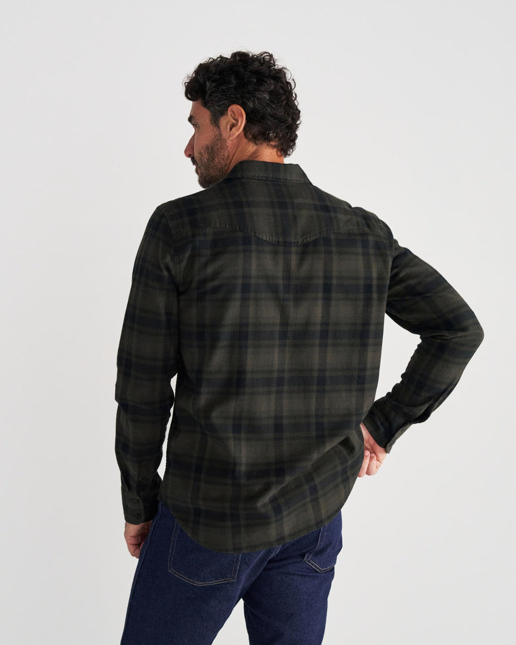 Man standing with his back to the camera, wearing a dark green and black plaid shirt and blue jeans against a plain white background.