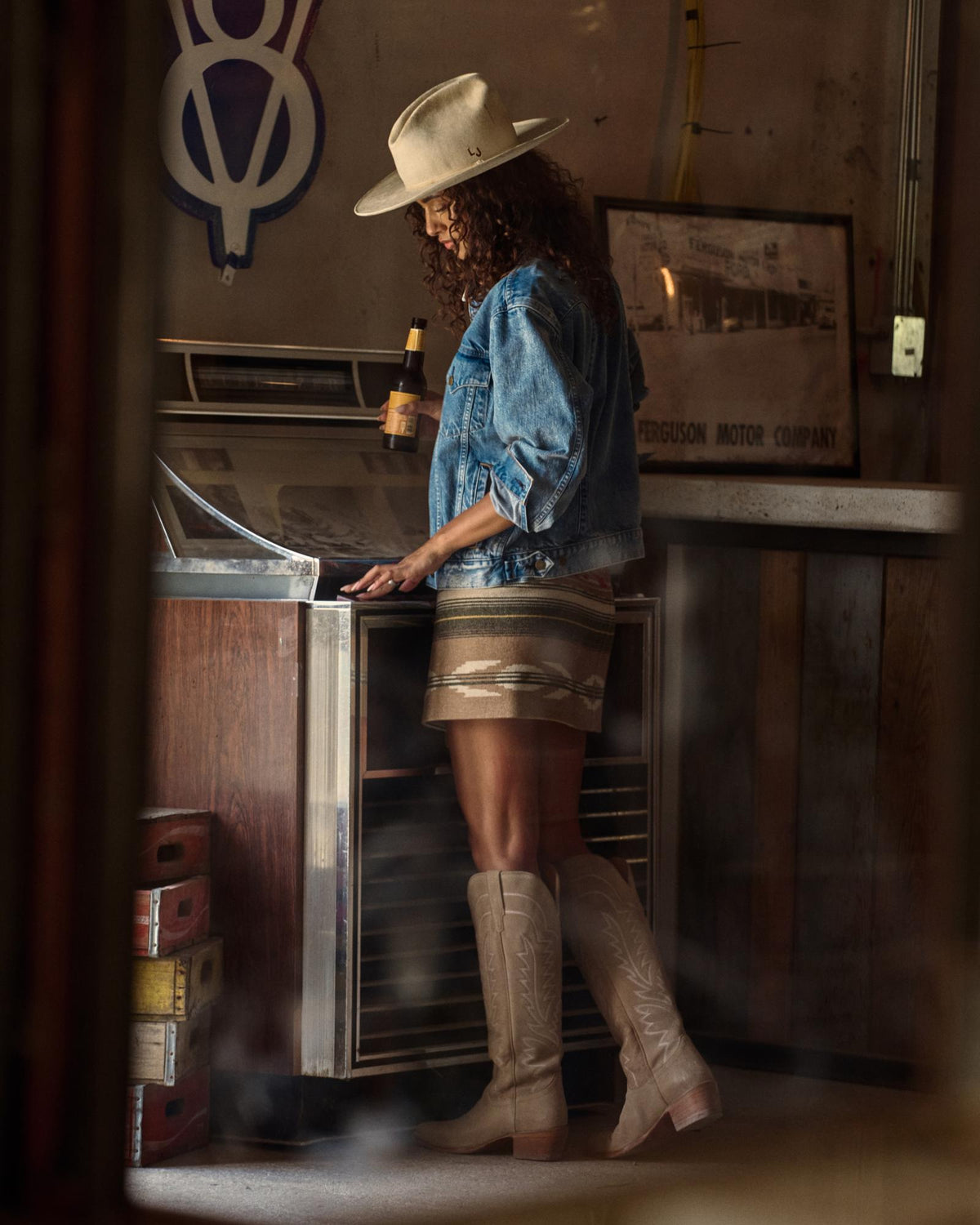 The Abby in Driftwood Suede. A woman in a hat, denim jacket, mini skirt, and The Abby Suede cowboy boots stands at a retro cooler, holding a bottle, in a rustic indoor setting.