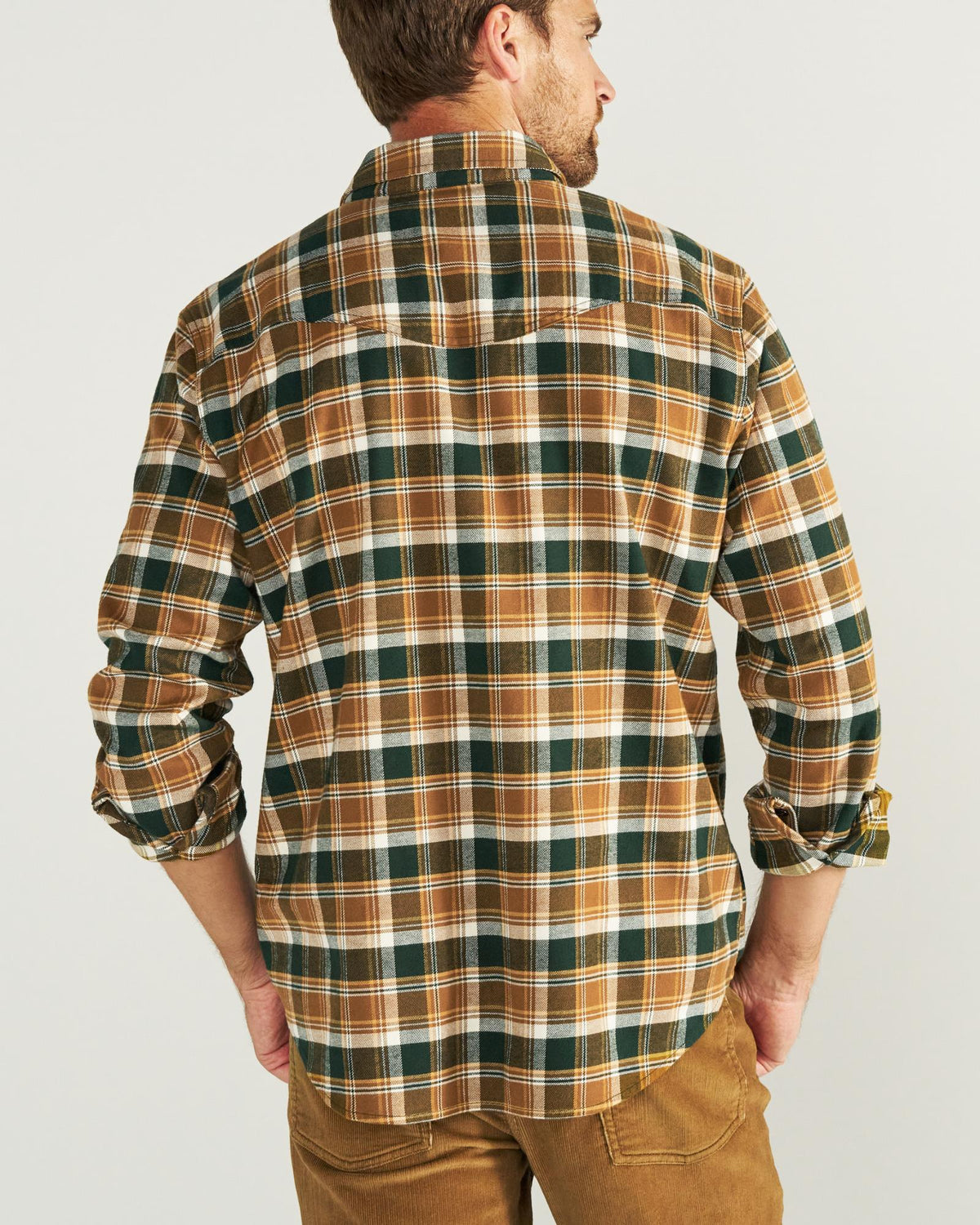 A man stands facing away, wearing a brown, green, and white plaid long-sleeve shirt with brown pants against a plain light background.
