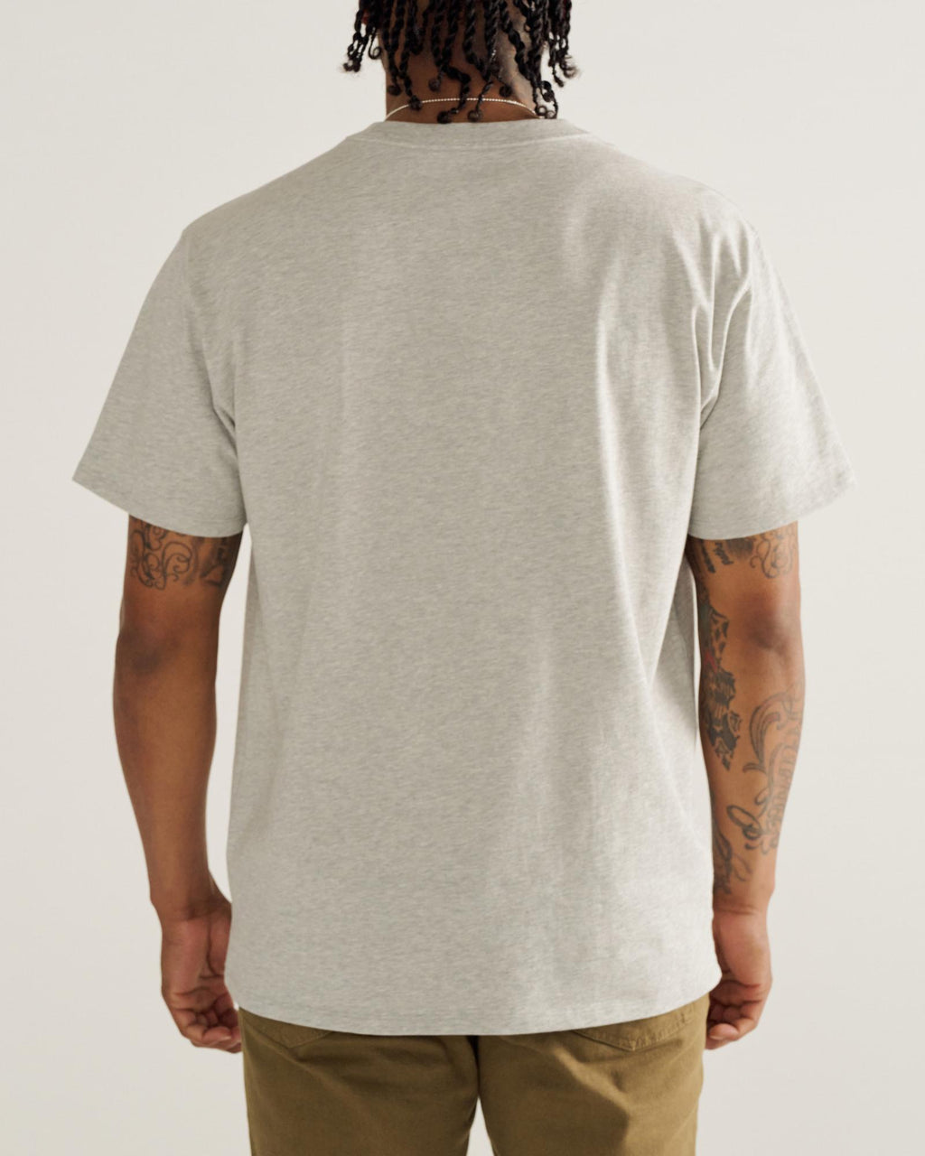 A person with tattoos on both arms is standing facing away, wearing a light gray T-shirt and olive green pants against a plain background.
