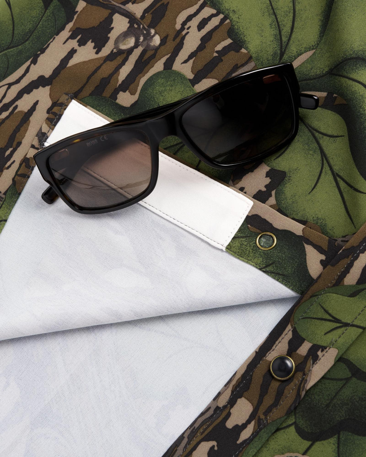 Black rectangular sunglasses rest on a camouflage-patterned shirt with green leaf and brown stripe designs, partially folded to reveal a white interior and metal snap buttons.