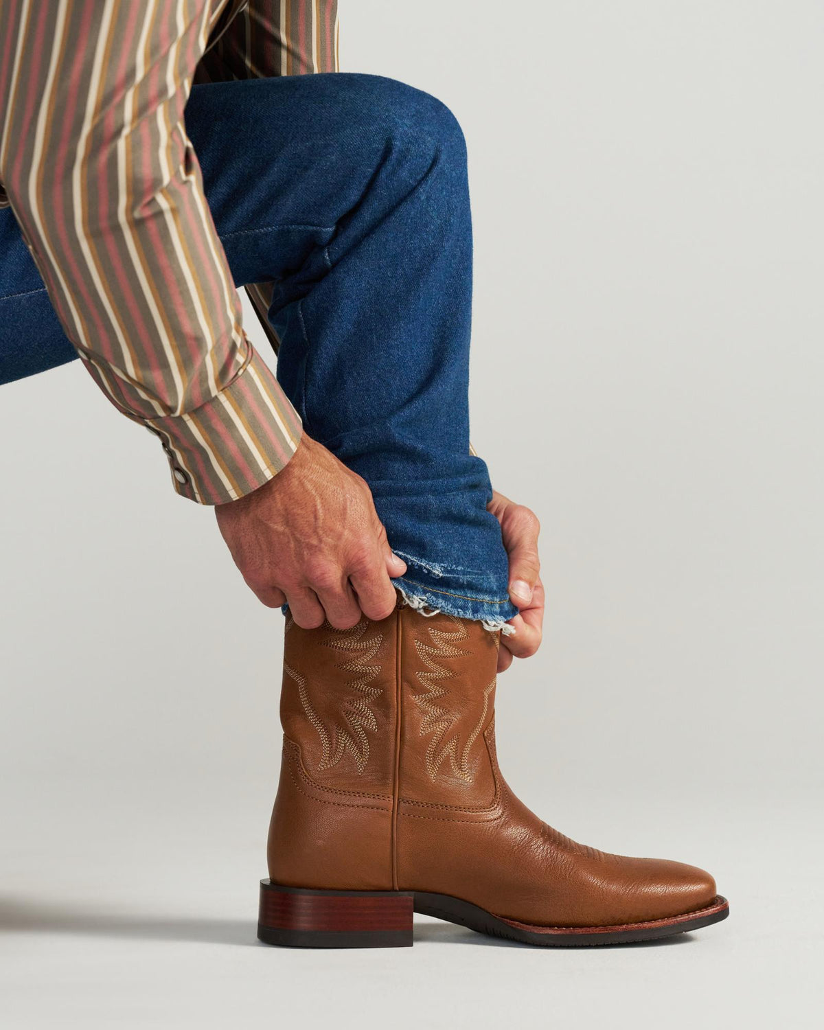 The Dusty in Mesa Goat. Person wearing a striped shirt and blue jeans, pulling a pant leg over a tan leather cowboy boot with decorative stitching, ready to explore the rugged trails of Mesa.