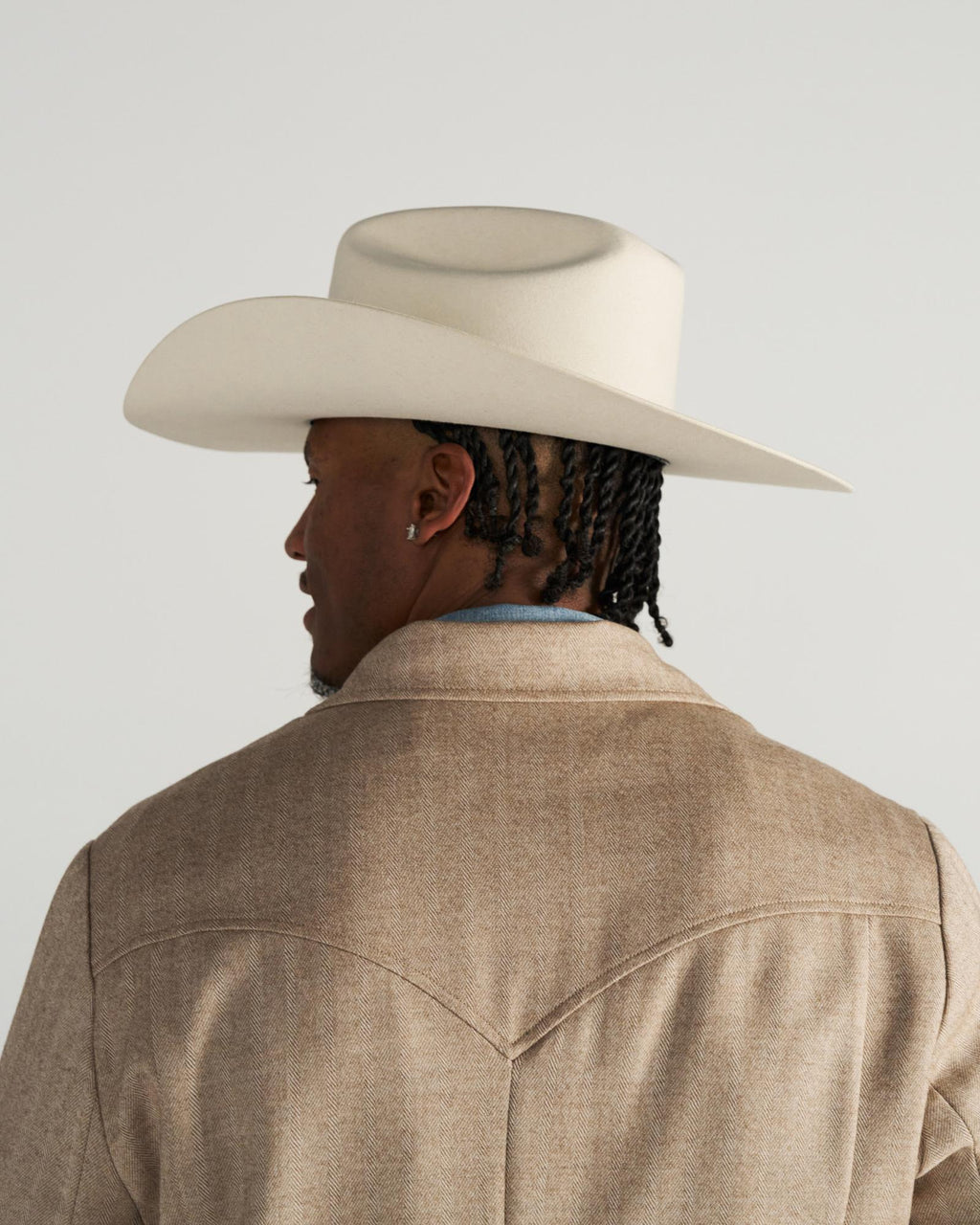 A person with braided hair, wearing a light-colored cowboy hat and a tan jacket, is shown from behind against a plain background.