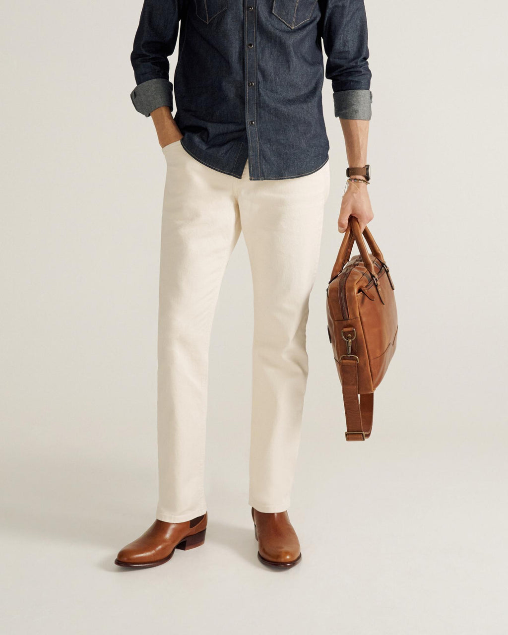 A person wearing a dark denim shirt, white pants, and brown boots holds a brown leather bag in their left hand.