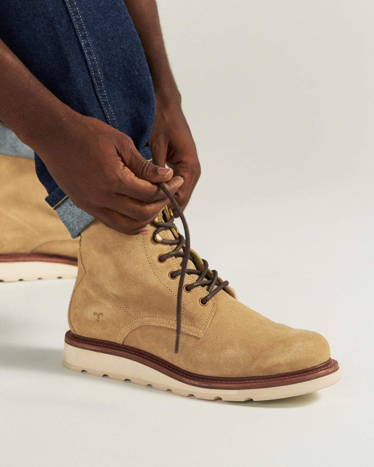 The Knox Lace Up Shearling in Sandstone Roughout. Person tying the brown laces of The Knox Lace Up Shearling Roughout tan suede boot with a white sole, wearing blue jeans, against a neutral background.
