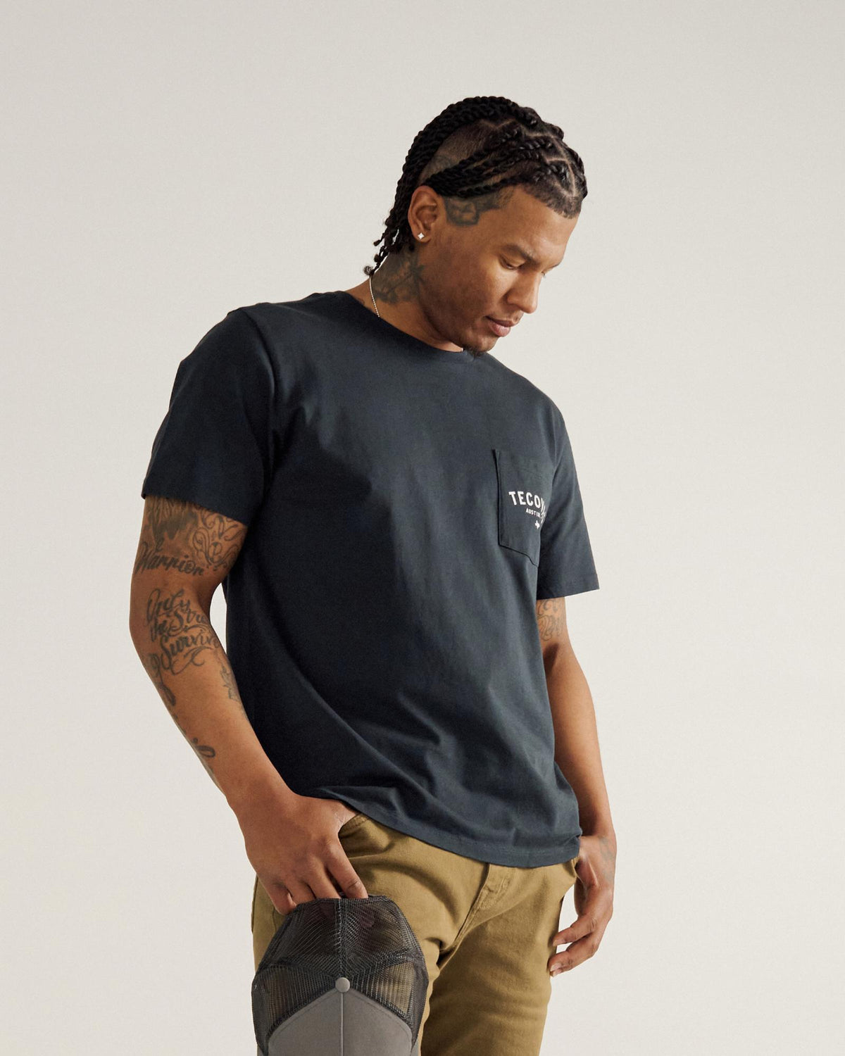 A man with braided hair and visible tattoos stands looking down, wearing a dark T-shirt with a chest pocket and khaki pants, holding a gray cap in one hand against a plain background.