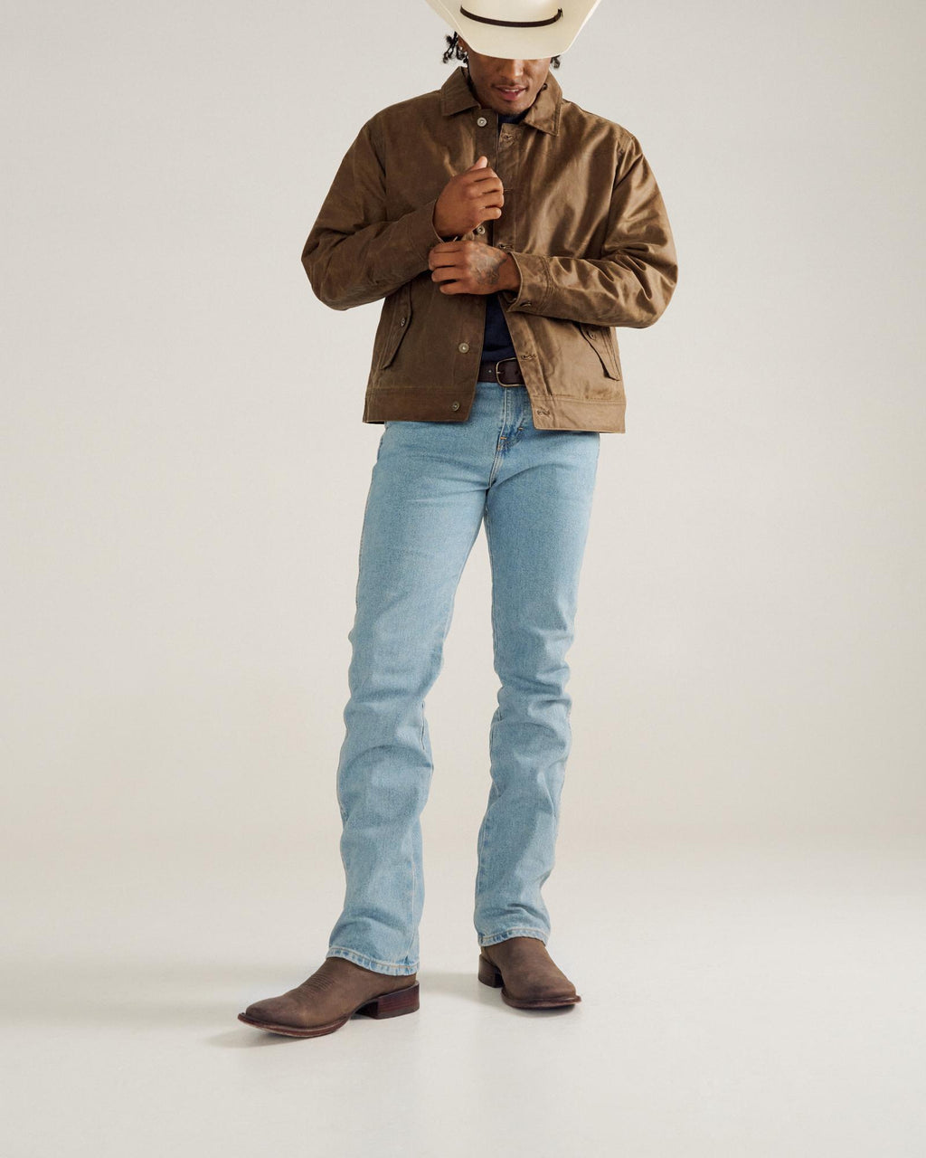 Person wearing a brown jacket, light blue jeans, brown cowboy boots, and a white cowboy hat against a plain background. Face is partially obscured by the hat.