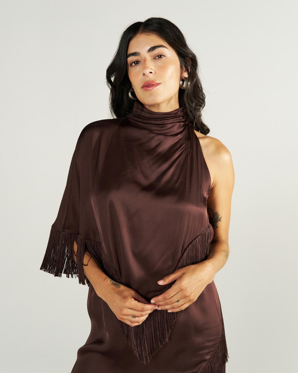 Woman with long dark hair wearing a shiny, dark brown, asymmetrical dress with fringe detail, posing against a plain light background.