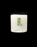 A white candle in a clear glass holder with a green illustration of a cowboy boot on the front.